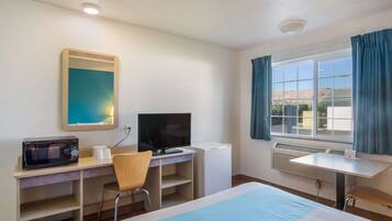 A hotel room with a bed, a desk with a computer, a chair, a microwave, a TV, and a window with curtains.
