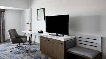 A hotel room with a desk, chair, TV, and a small sofa.