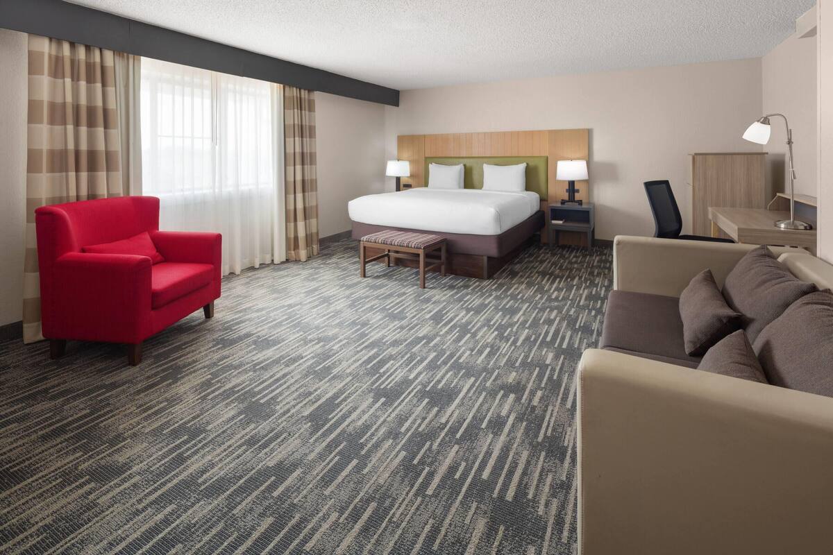 A hotel room with a large bed, a red armchair, a small table, a sofa, and a desk with a lamp.