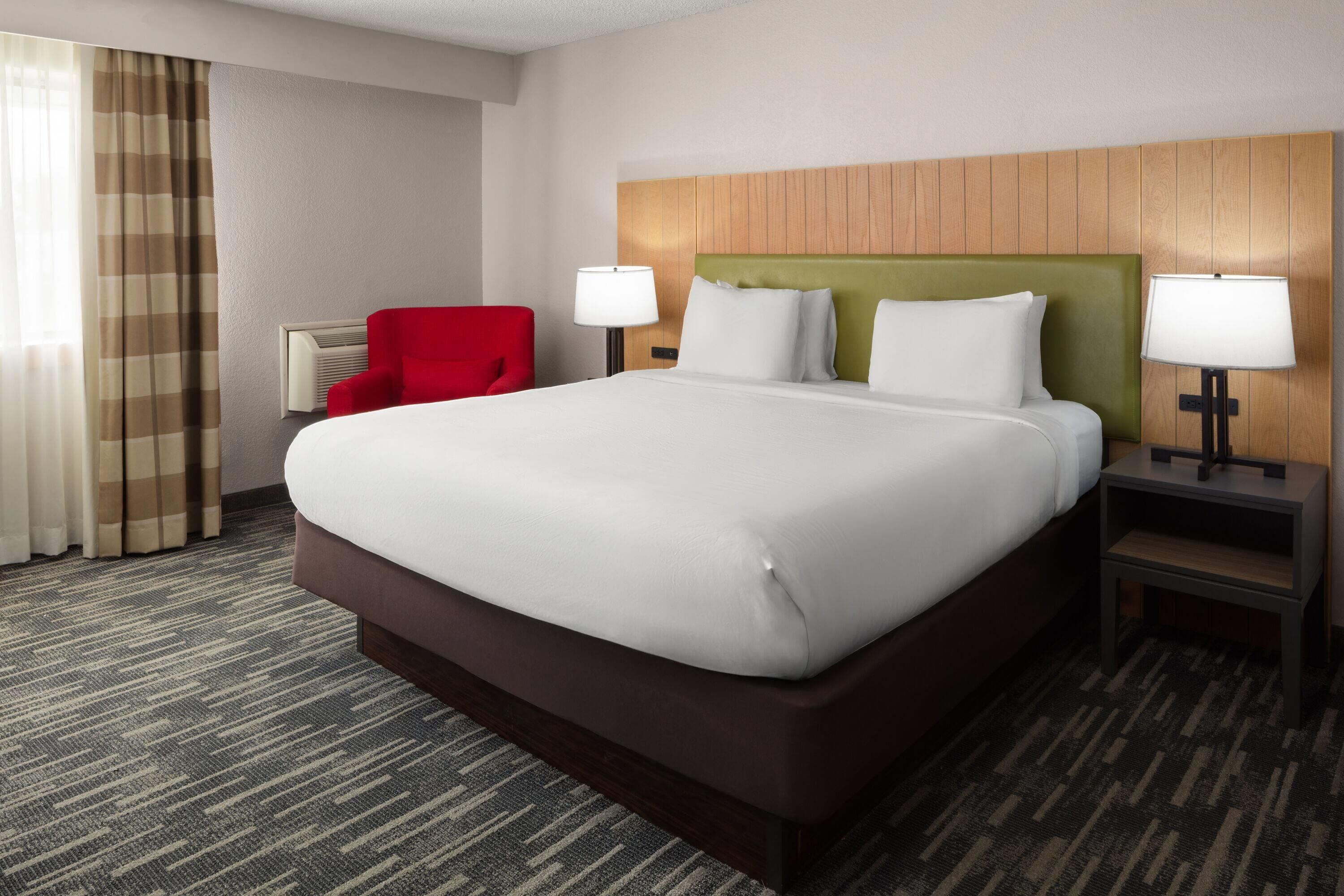 A hotel room with a large bed, two bedside tables with lamps, a red armchair, and a window with curtains.