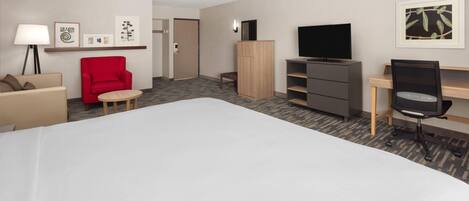 A hotel room with a large bed, a red armchair, a desk with a chair, a TV, and a wardrobe.