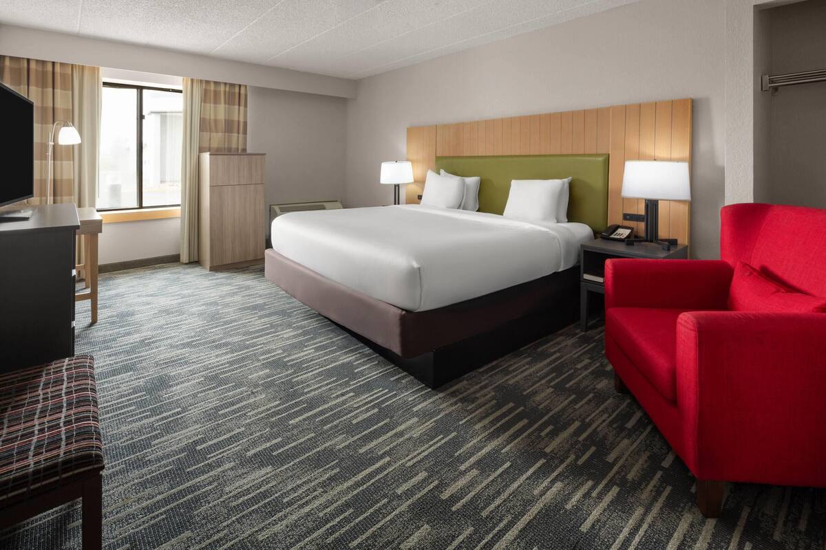 A hotel room with a large bed, a desk with a TV, a red armchair, and a window with curtains.