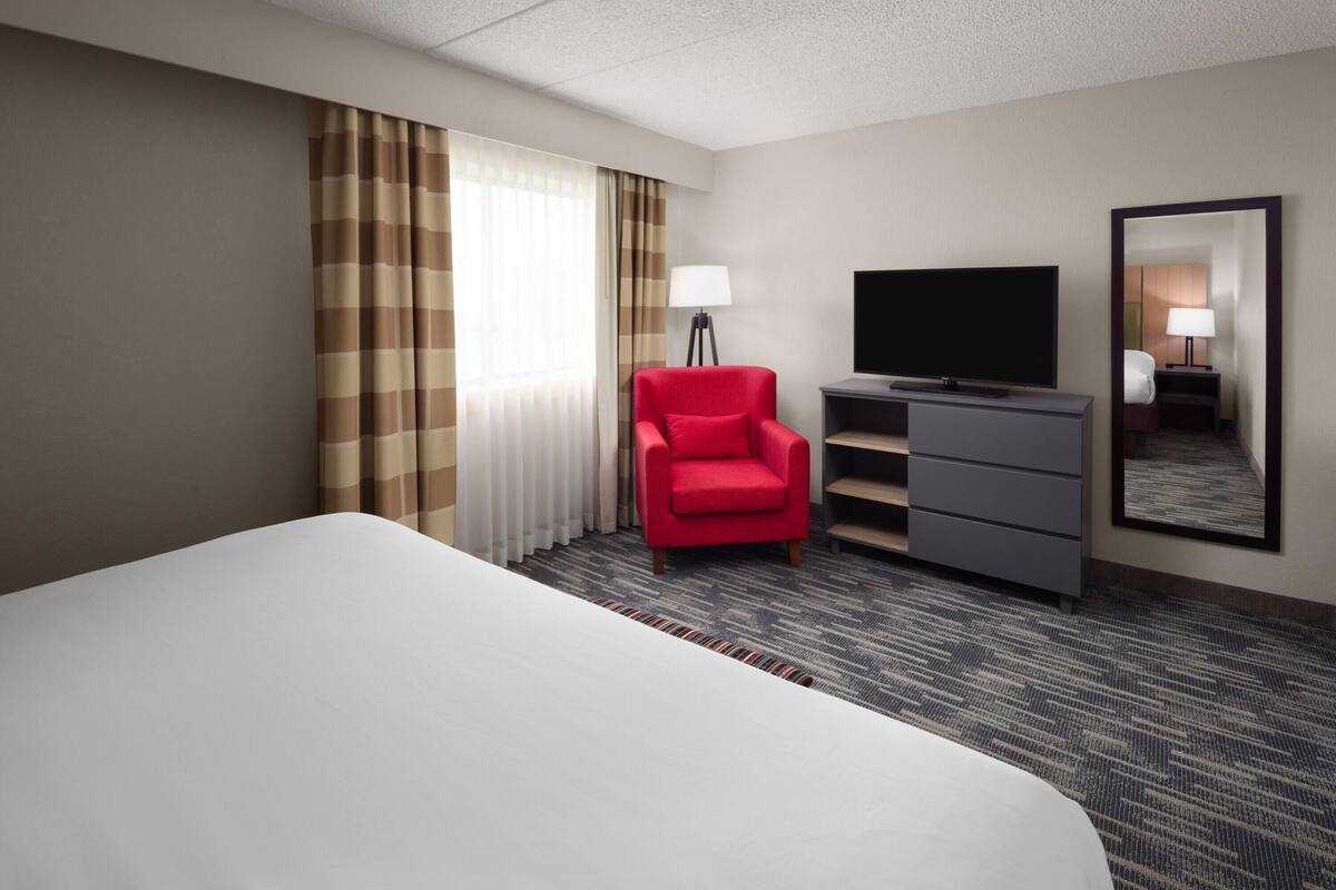 A hotel room with a large bed, a red armchair, a TV, a dresser, and a mirror.
