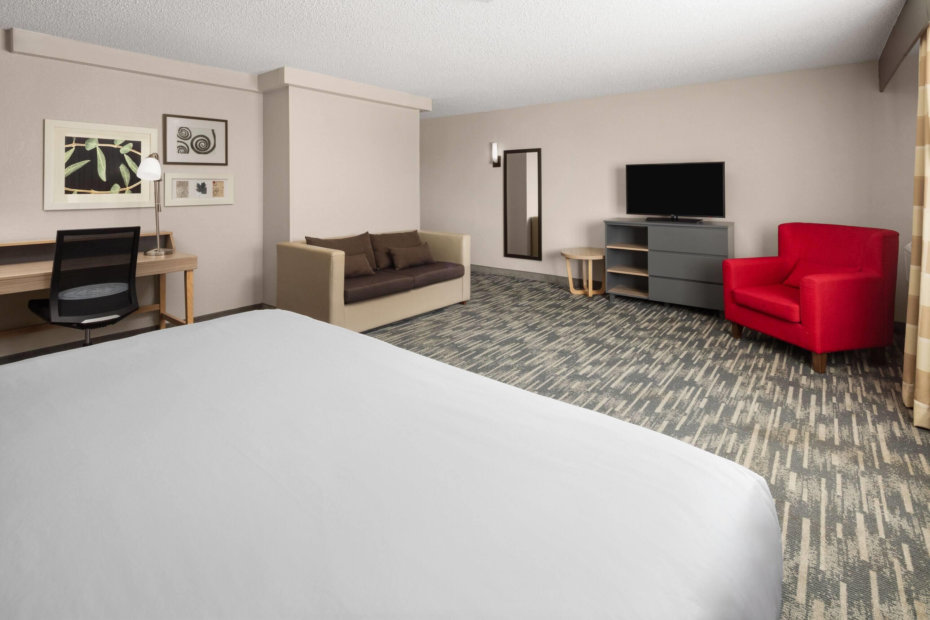 A hotel room with a large bed, a desk with a chair, a sofa, a red armchair, a TV, and a small side table.