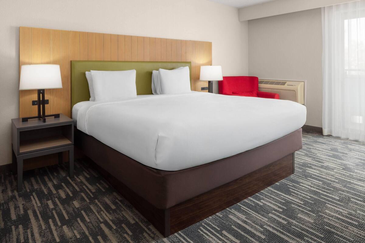 A hotel room with a large bed, two bedside tables with lamps, a red chair, and a wall-mounted air conditioner.