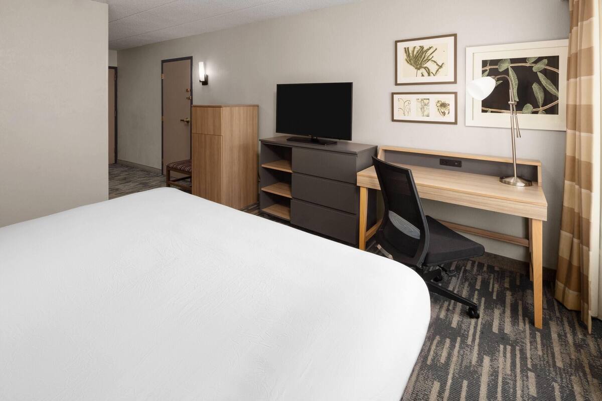 A hotel room with a bed, a desk with a computer, a television, and a lamp. There are framed botanical prints on the wall.