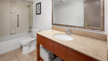 Un baño con inodoro blanco, lavabo con tocador de madera, espejo grande y zona de ducha.