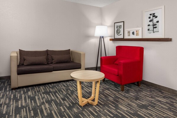 Habitación de hotel con sofá color beige, un sillón rojo, una mesita de madera y una lámpara de pie. En la pared hay cuadros enmarcados.