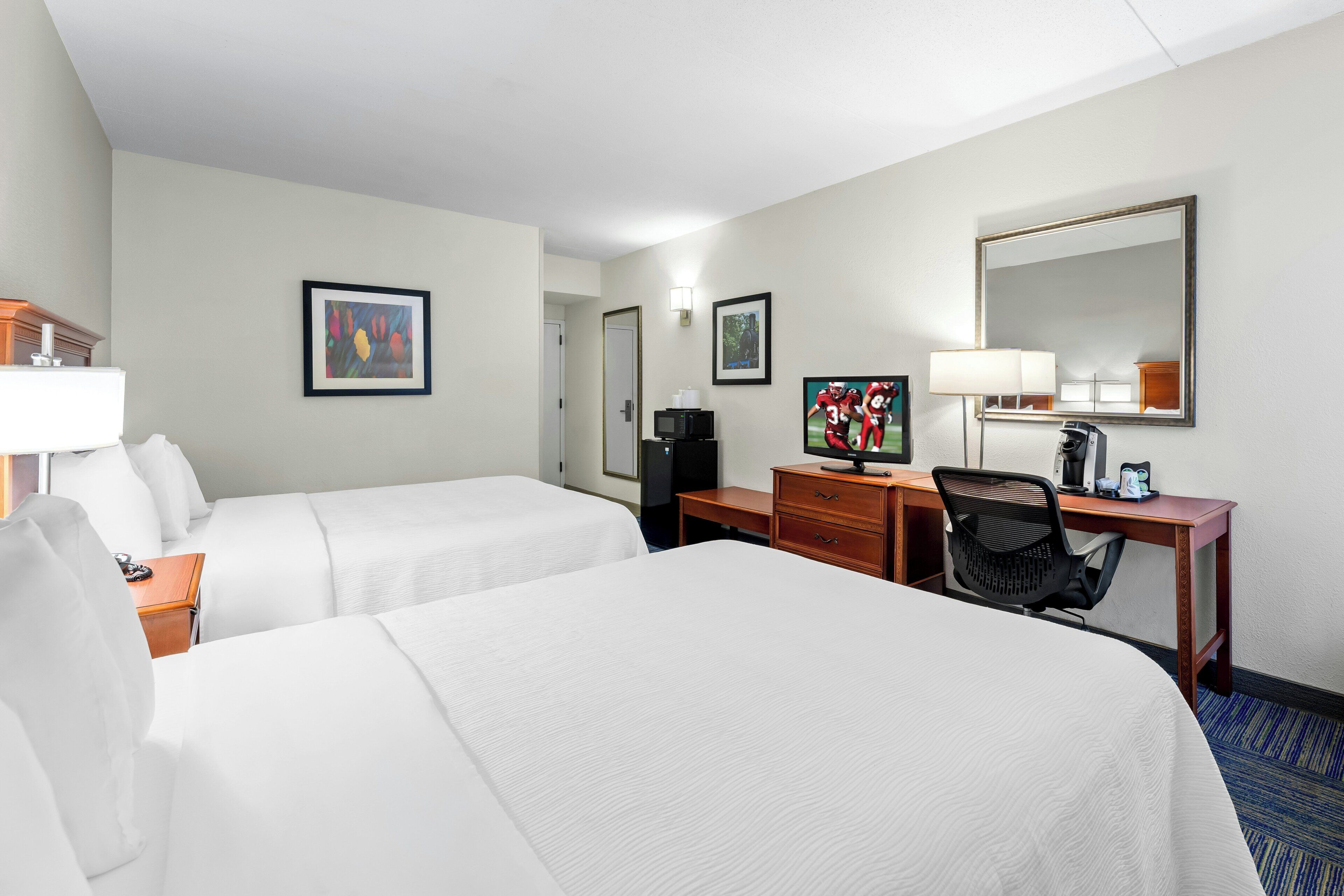 A hotel room with two beds, a desk, a chair, a television, a coffee maker, and framed artwork on the walls.