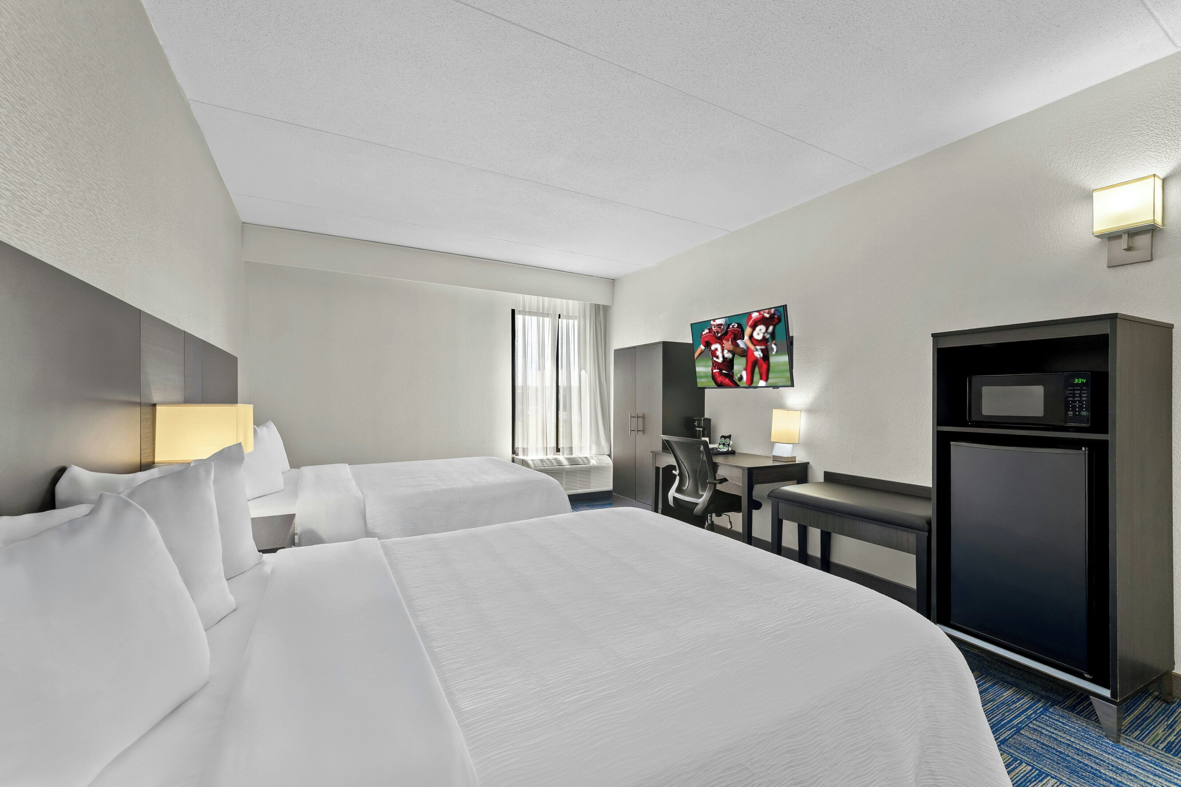 Quarto de hotel com duas camas, uma escrivaninha, televisão, micro-ondas e janela com cortinas.