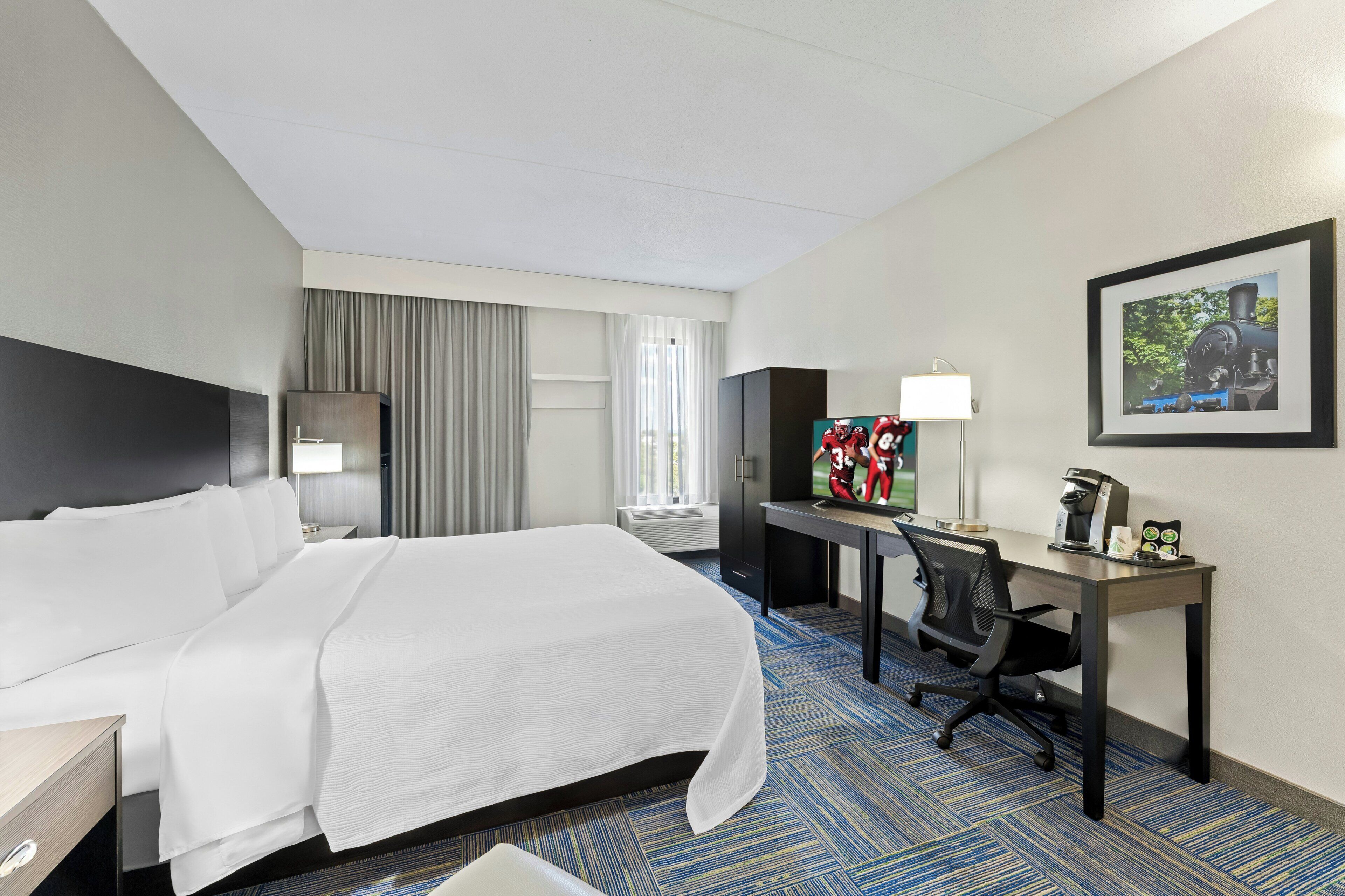 A hotel room with a bed, desk, chair, lamp, and a framed picture on the wall.
