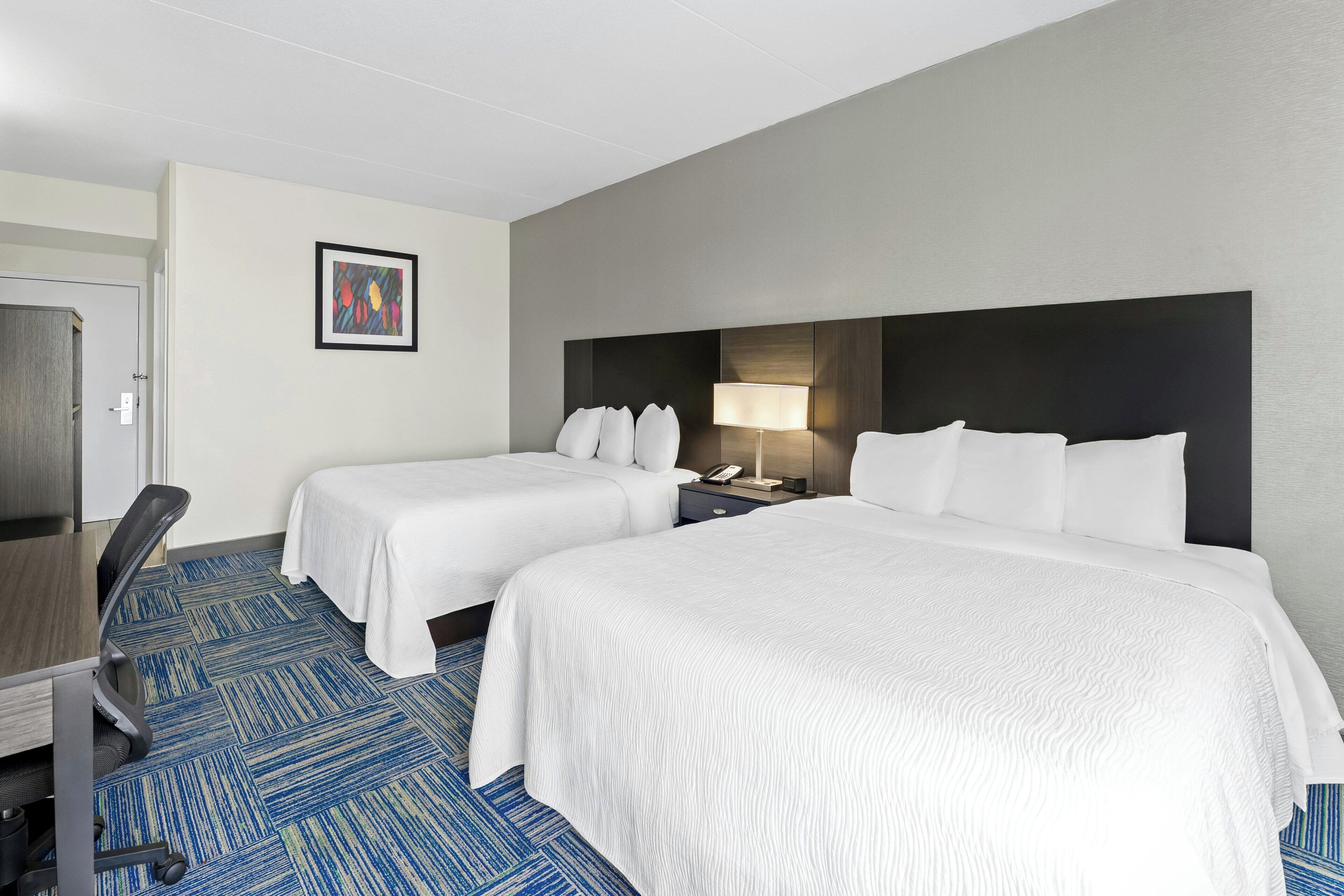 A hotel room with two beds, a desk, a chair, a lamp, and a framed picture on the wall.