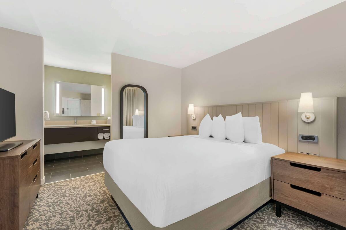 A hotel room with a large bed, a wooden nightstand, a bedside table with a lamp, a mirror, a TV, and a bathroom visible in the background.