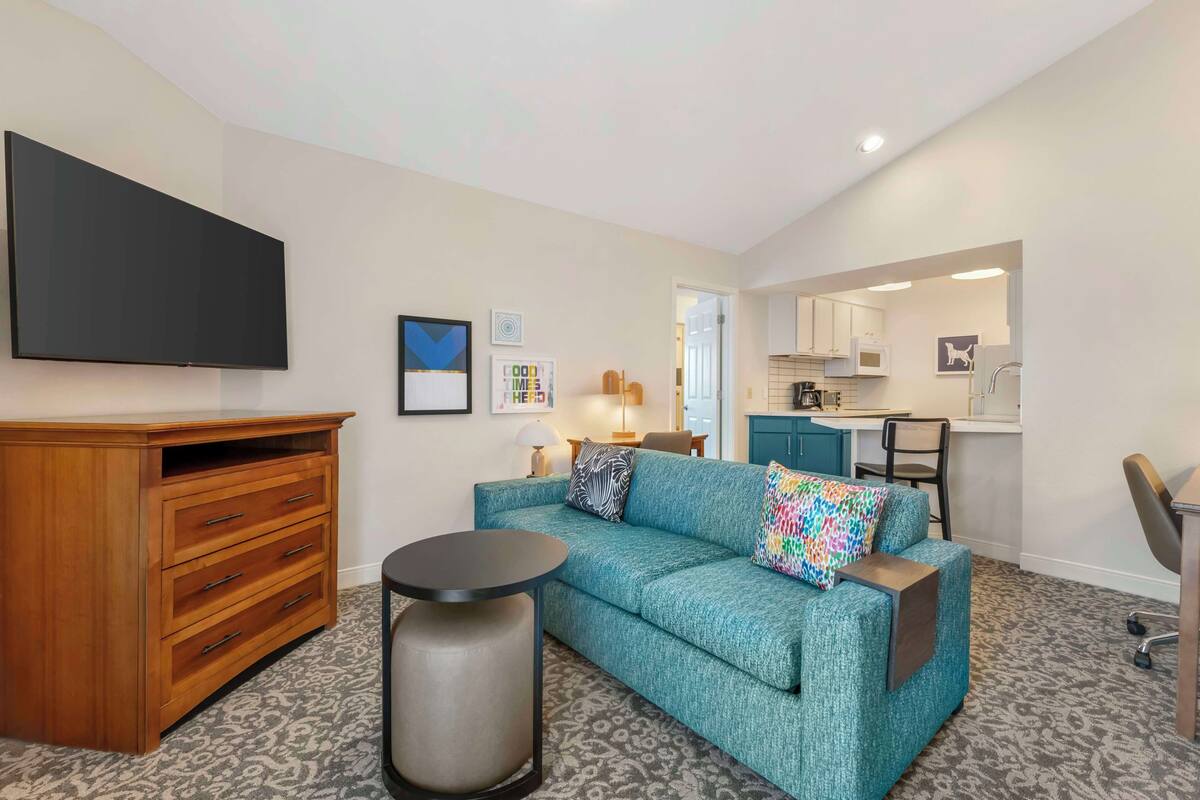 A compact living space with a sofa, a wooden dresser, a round table, and a kitchenette in the background.