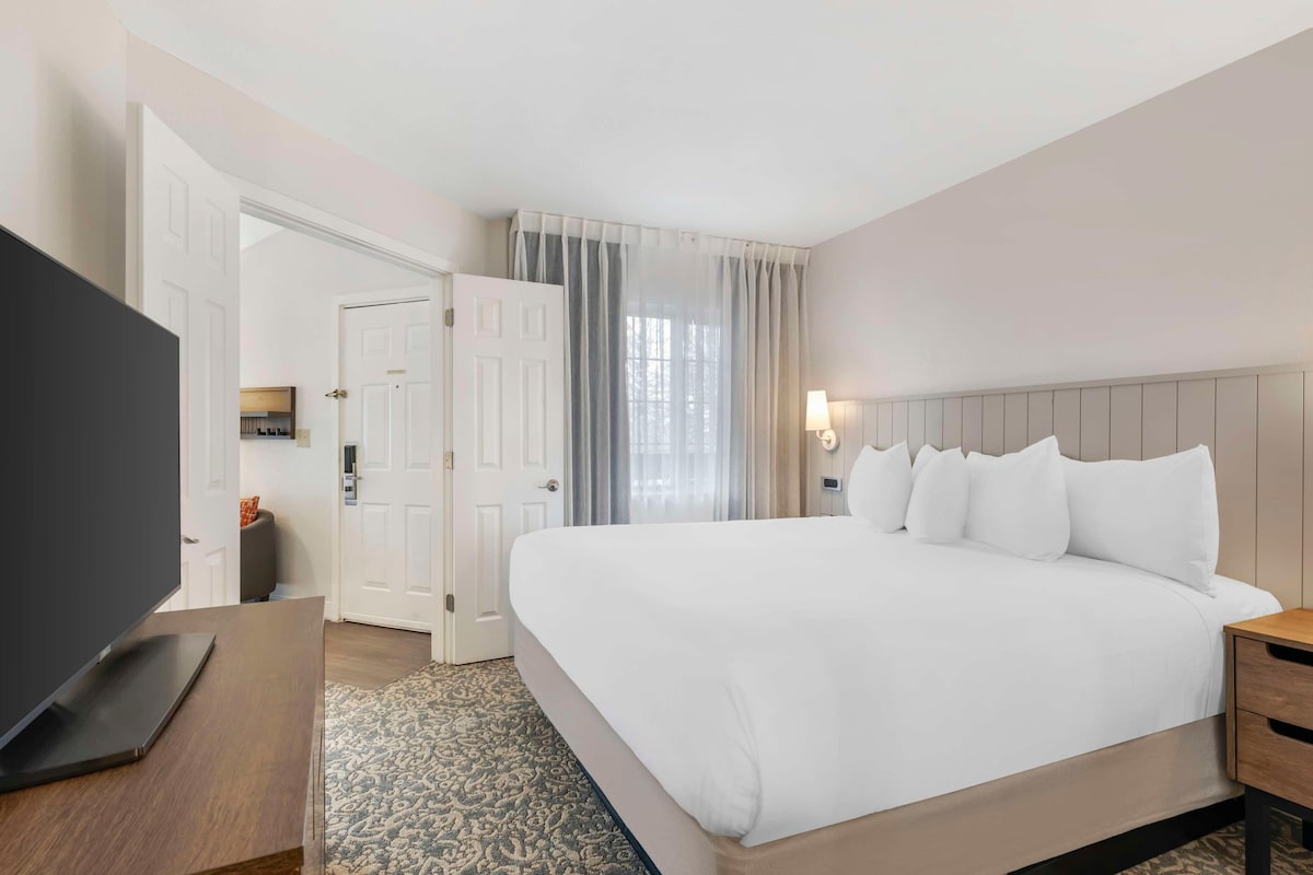 Una habitación de hotel con una cama grande, un televisor sobre una mesa de madera, una lámpara de noche y una puerta que da a otra habitación.
