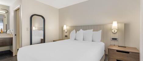 Habitación de hotel con una cama grande, dos mesitas de noche con lámparas, un espejo y un baño visible a través de una puerta abierta.