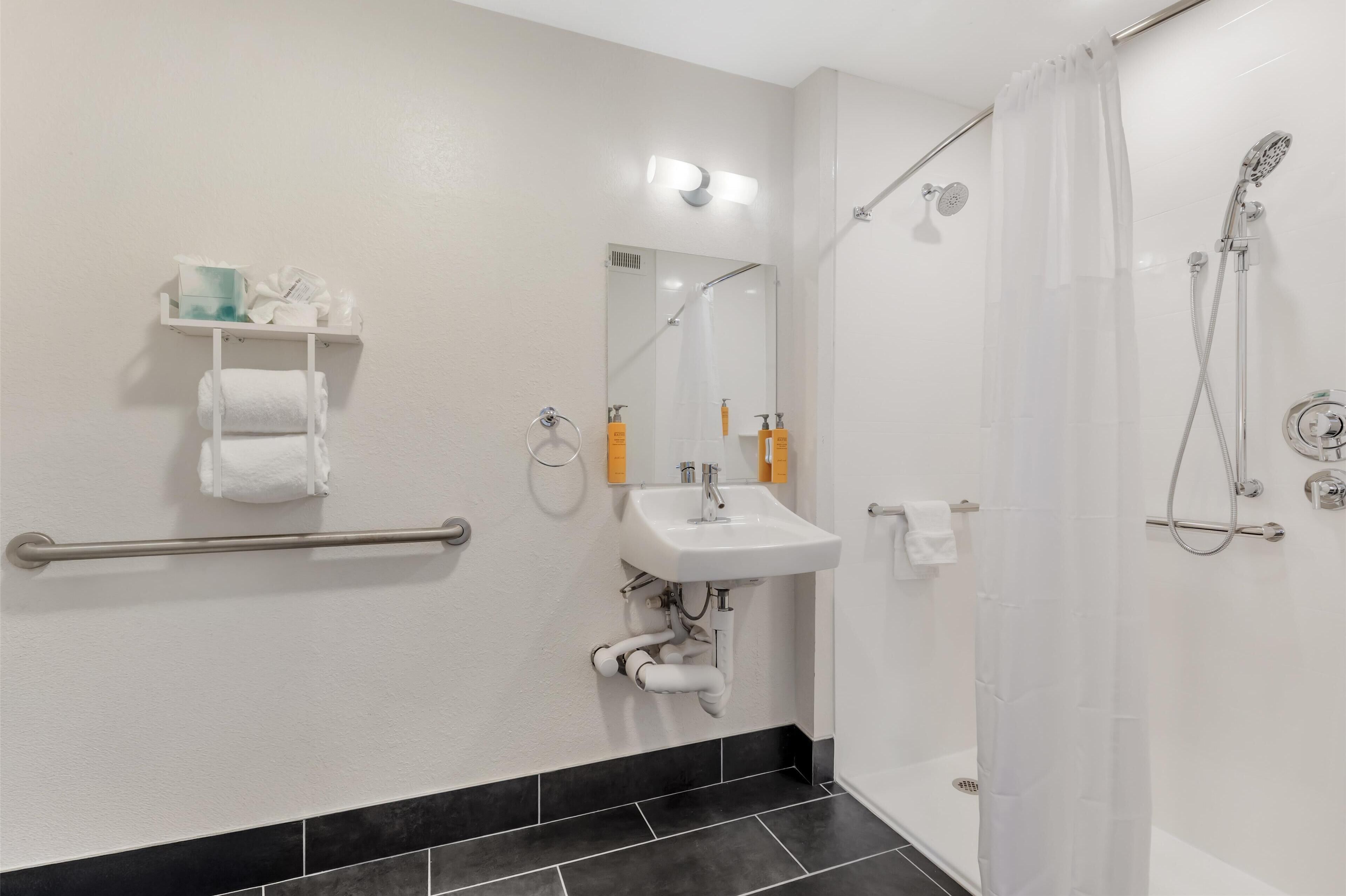Un baño moderno con lavamanos, espejo y ducha blancos. Incluye un perchero y estante con artículos de aseo.