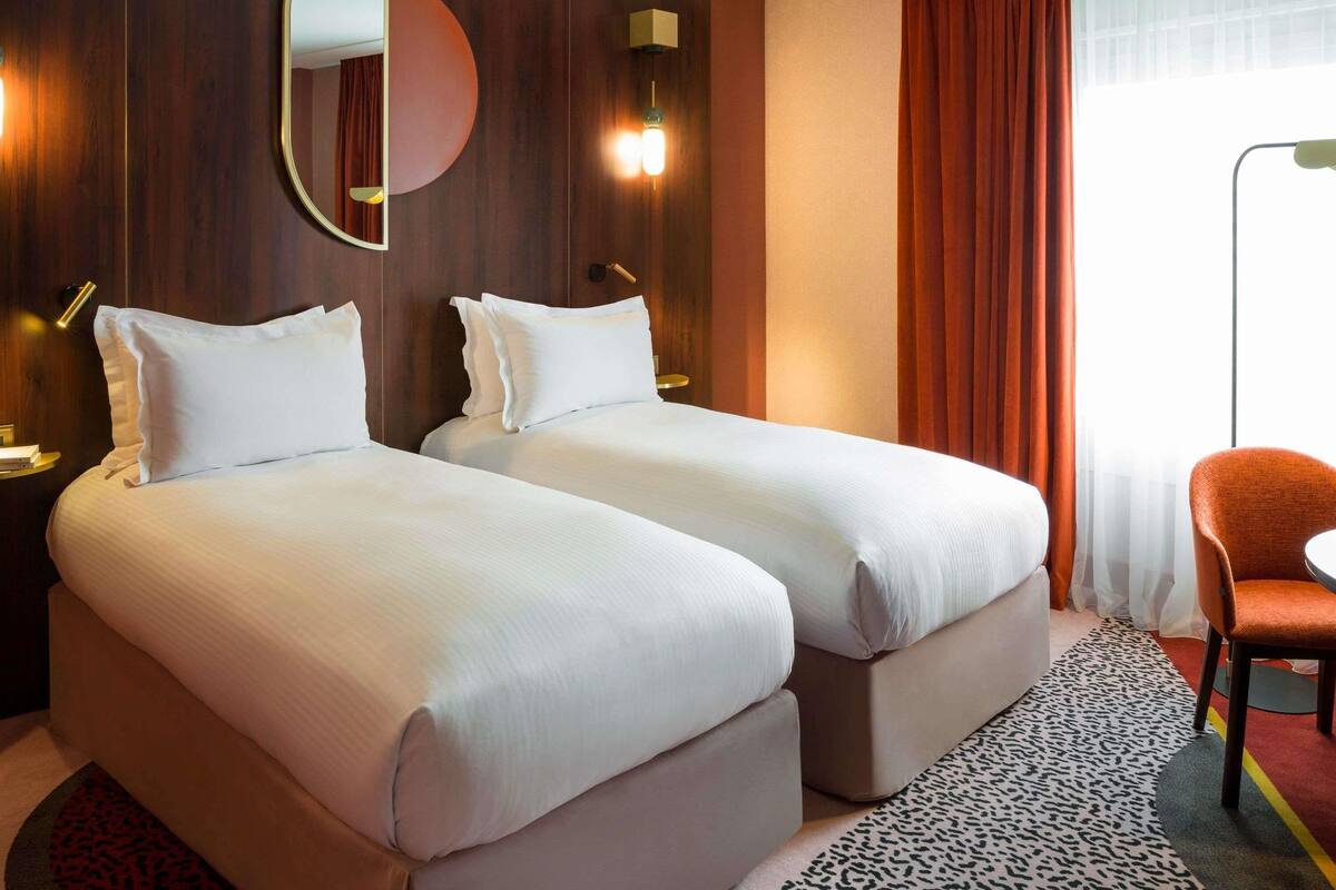 A hotel room with two beds, a round mirror, wall sconces, a chair, and a window with curtains.