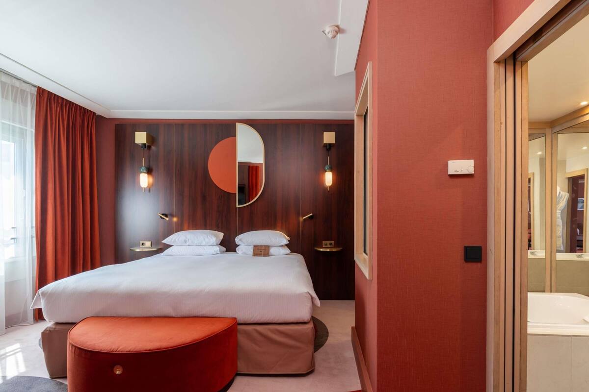 A hotel room with a bed, two bedside tables, a mirror, and a red ottoman.
