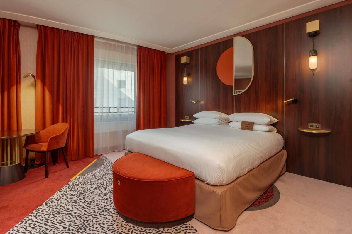 A hotel room with a bed, a desk, a chair, a round ottoman, a mirror, and a window with curtains.