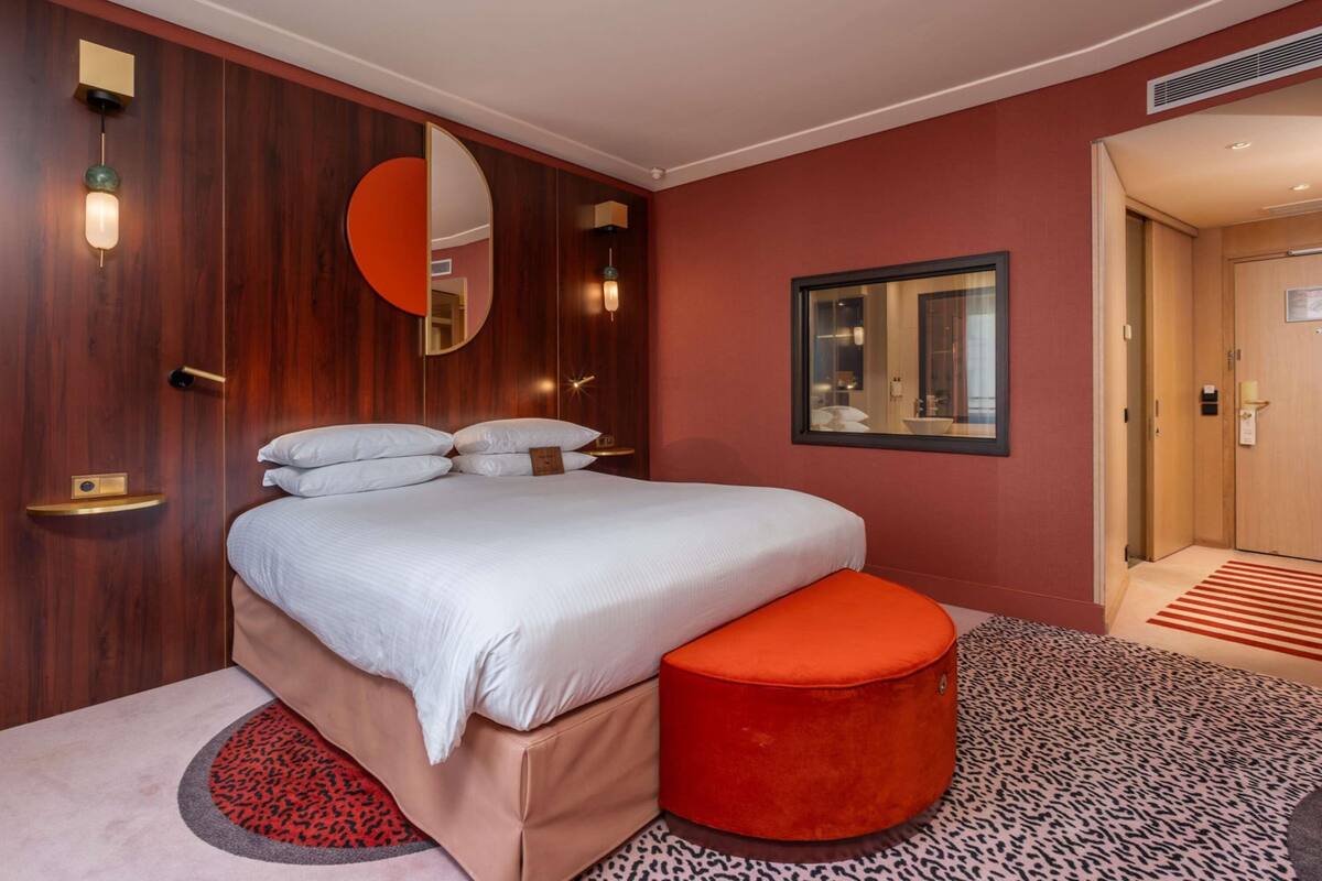 A hotel room with a bed, a round ottoman, a mirror, and a door.