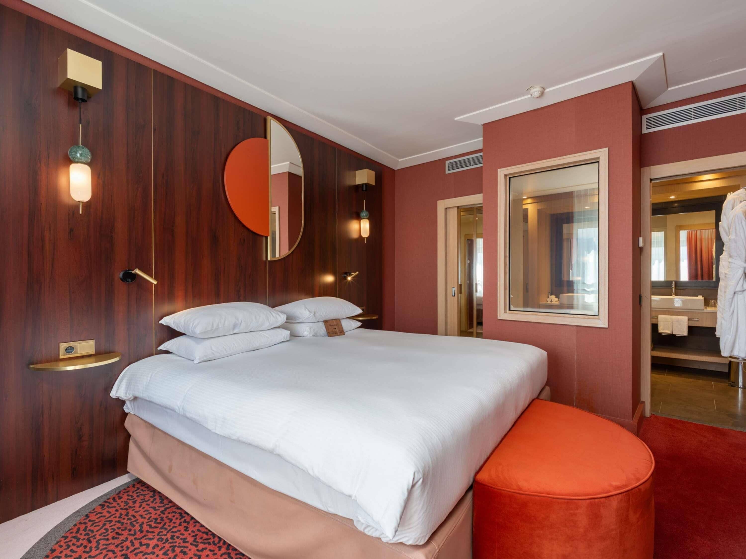 Una habitación de hotel con cama, un espejo redondo, un reposapiés rojo y un baño visible a través de una puerta abierta.