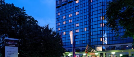 Un hotel moderno con fachada de vidrio, un cartel que enumera servicios como restaurante y bar, y coches estacionados frente a él.
