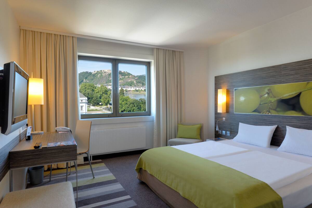 Habitación de hotel con cama, escritorio con silla, televisión y ventana con vista a una colina y edificios.