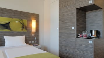 A modern hotel room with a bed, a nightstand, a wall-mounted shelf with mugs and a coffee pot, and a painting of a fruit.
