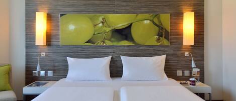 Habitación de hotel con una cama grande, dos almohadas blancas, un edredón verde, un cuadro colgado en la pared con uvas verdes y dos lámparas de noche.