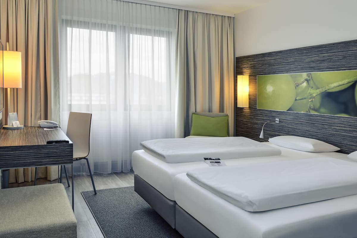 Habitación de hotel con cama, escritorio con lámpara, una silla y un ventanal con cortinas.