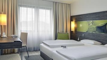Habitación de hotel con cama, escritorio con lámpara, una silla y un ventanal con cortinas.