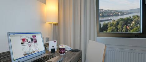 Habitación de hotel con una laptop sobre un escritorio, un teléfono, una taza y vista a un río y pueblo a través de una ventana.