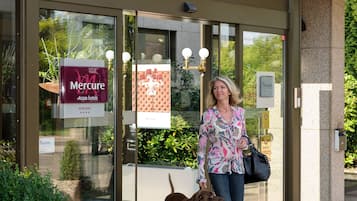 The exterior of the Mercure Hotel with a glass entrance, a large potted plant, and a dog walking in front.