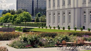 Un edificio de arquitectura clásica, un moderno edificio de cristal al fondo, y un jardín cuidado con un área de descanso.