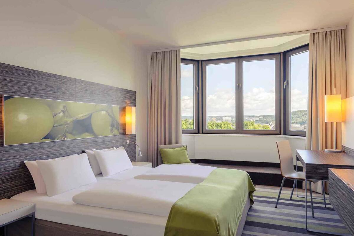 Habitación de hotel con una cama grande, un escritorio y vistas de la ciudad a través de amplias ventanas.