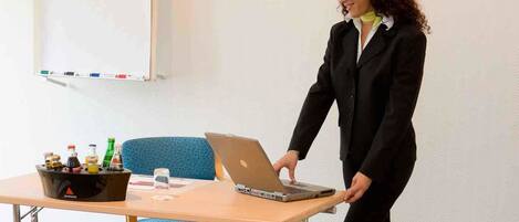 Una persona con traje negro de pie junto a una mesa con una computadora portátil, una botella de agua y un cuenco de fruta.