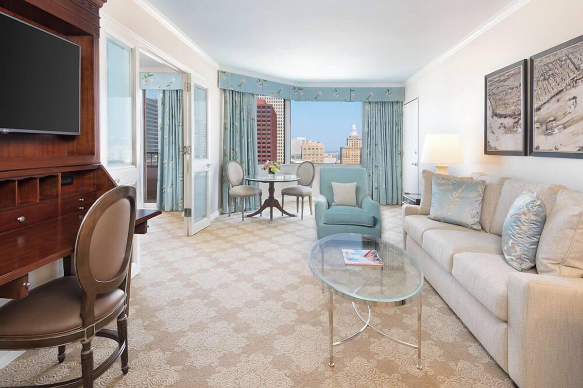 Quarto de hotel com área de refeições, sofá, televisão e vista para a paisagem urbana.