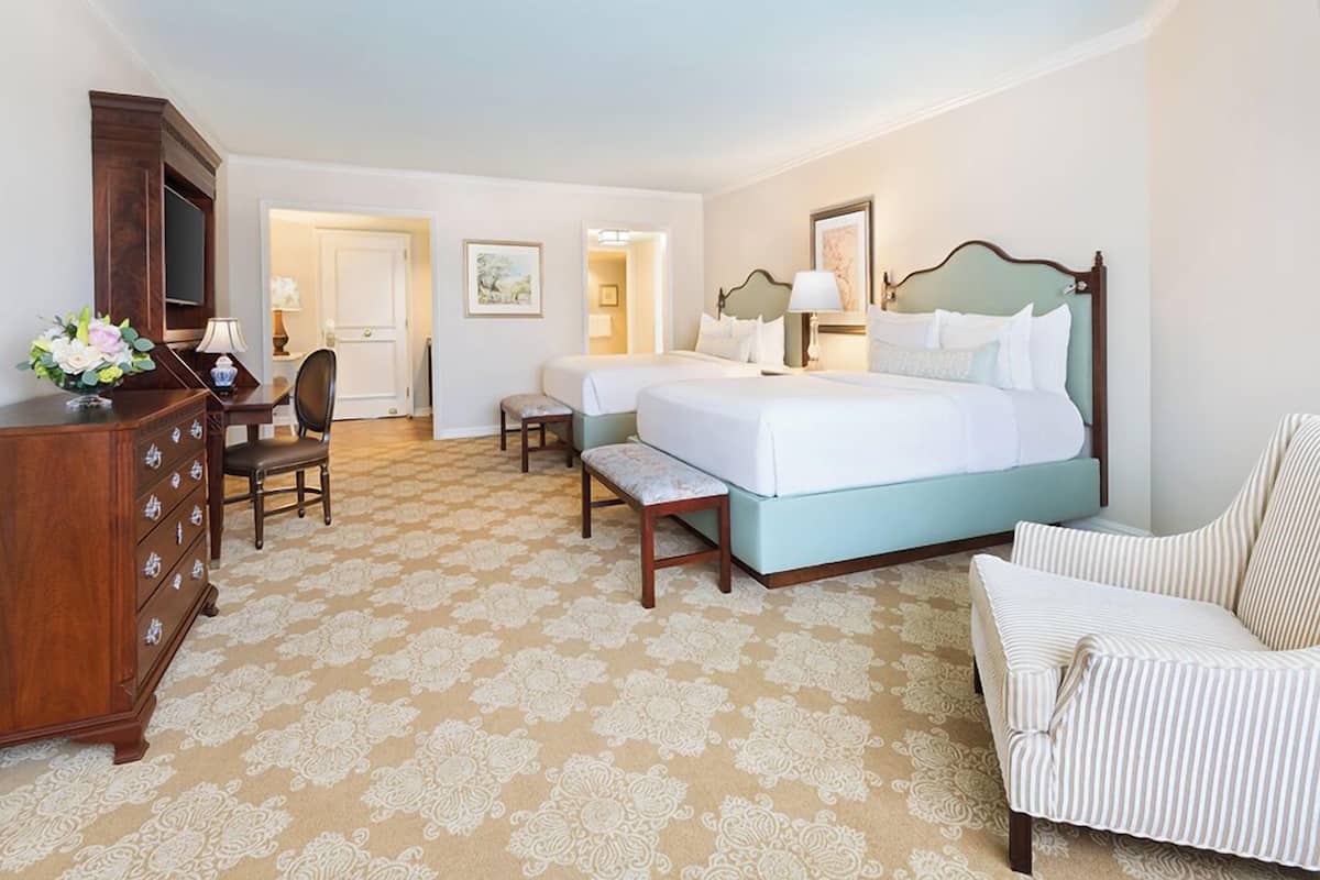 Quarto de hotel com duas camas, cômoda de madeira, uma cadeira e uma escrivaninha.