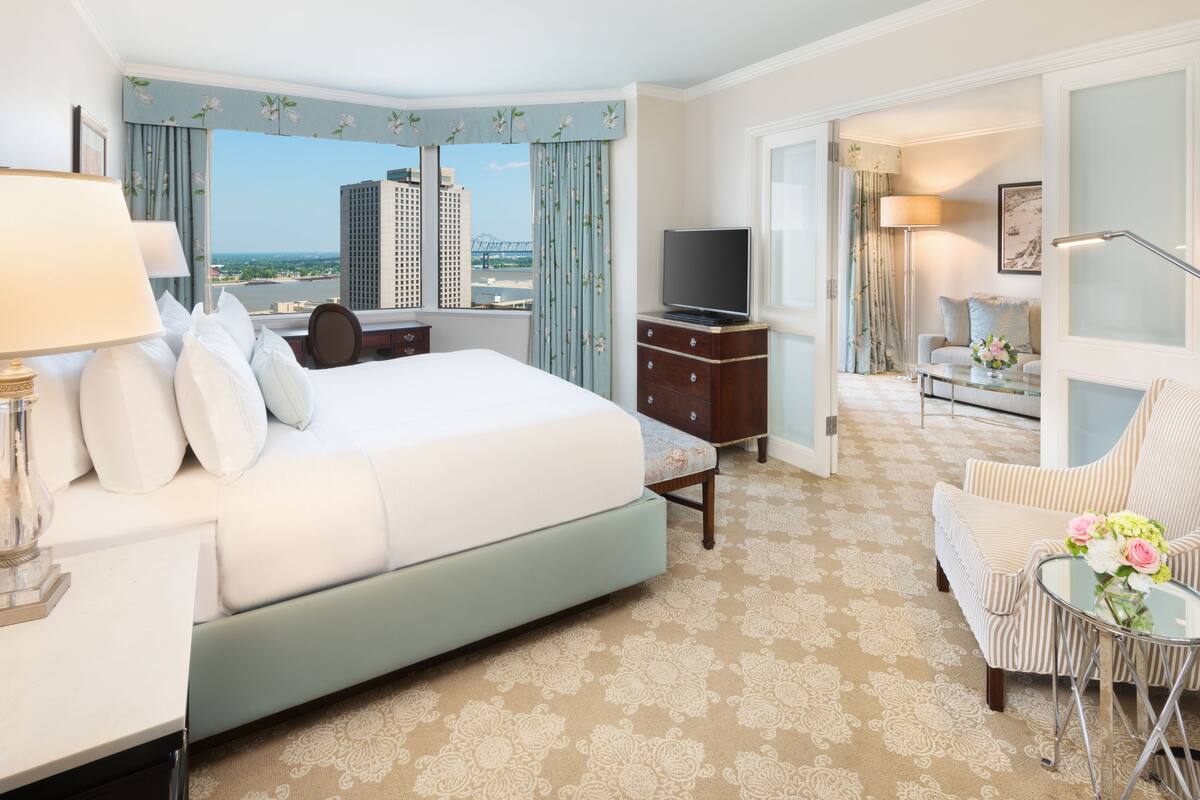 Quarto de hotel com uma cama grande, televisão, escrivaninha com luminária, cadeira e vista para a cidade.