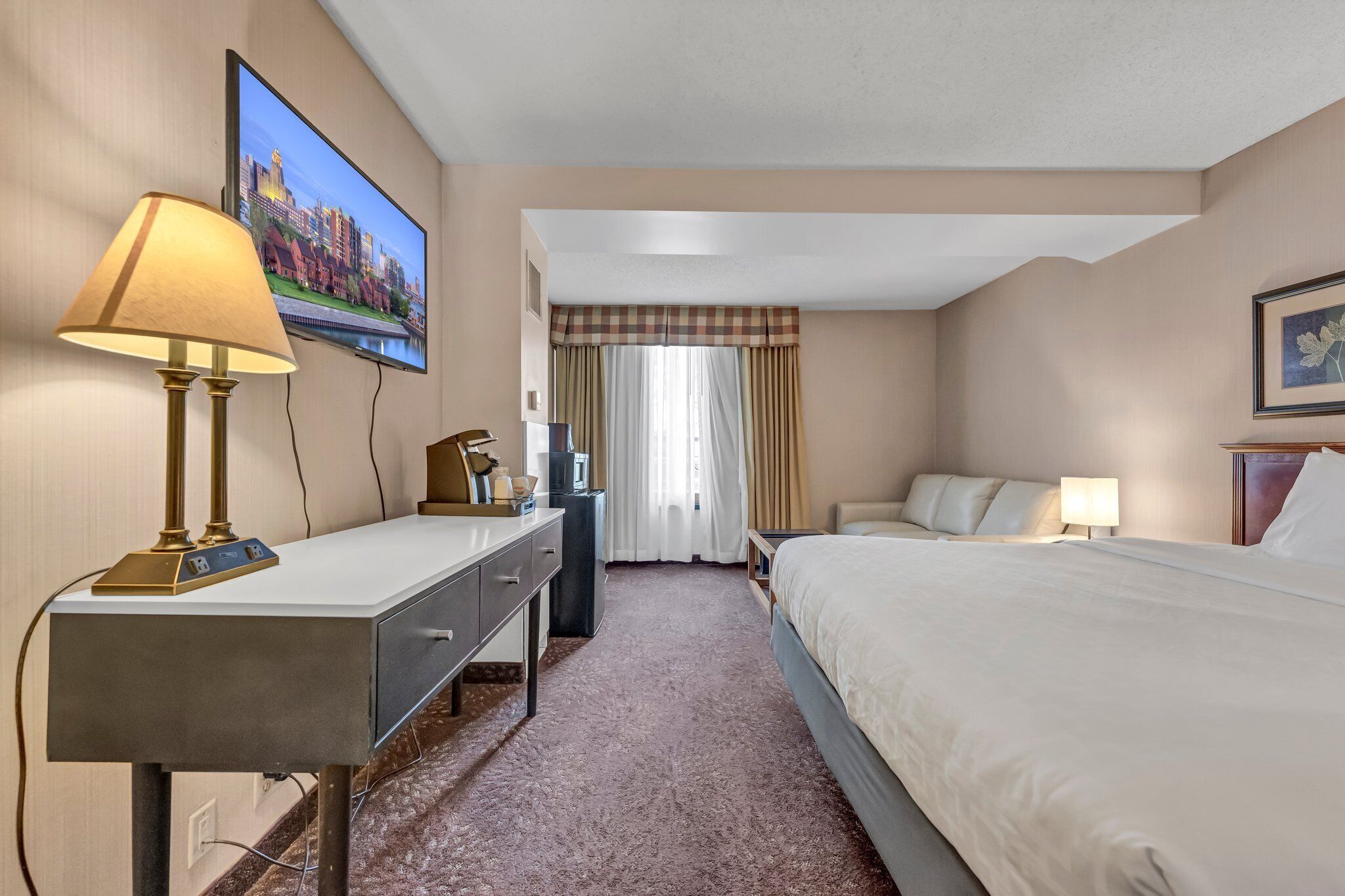 A hotel room with a bed, a desk with a lamp, a television, and a window with curtains.