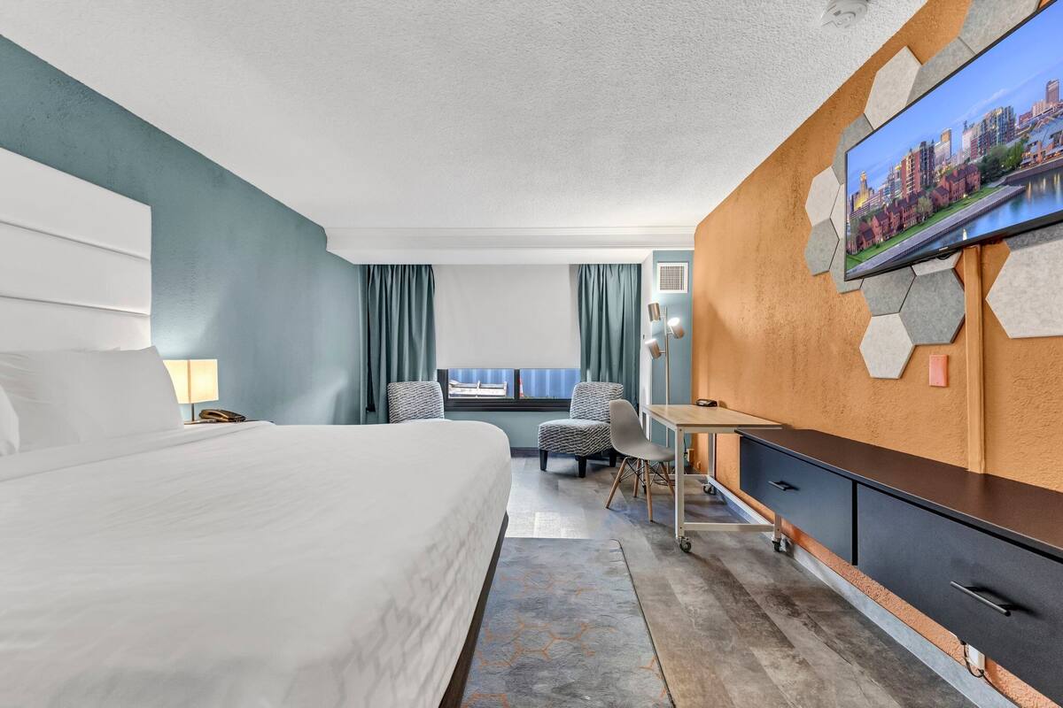 A hotel room with a large bed, a desk with a chair, a television mounted on the wall, and a window with curtains.