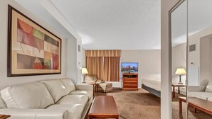 A hotel room with a beige sofa, a wooden coffee table, a TV, and a bed with a wooden headboard.