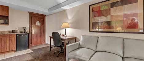 Habitación de hotel con kitchenette, un sofá, un escritorio con lámpara y un cuadro abstracto grande en la pared.