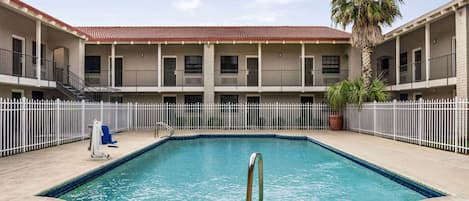 A pool area with a rectangular pool, a palm tree, and a building with balconies and a red-tiled roof.