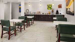 A dining area with green upholstered chairs and white tables, a kitchen counter with various food items, and artwork on the walls. - La Quinta Inn by Wyndham Victoria (Victoria)