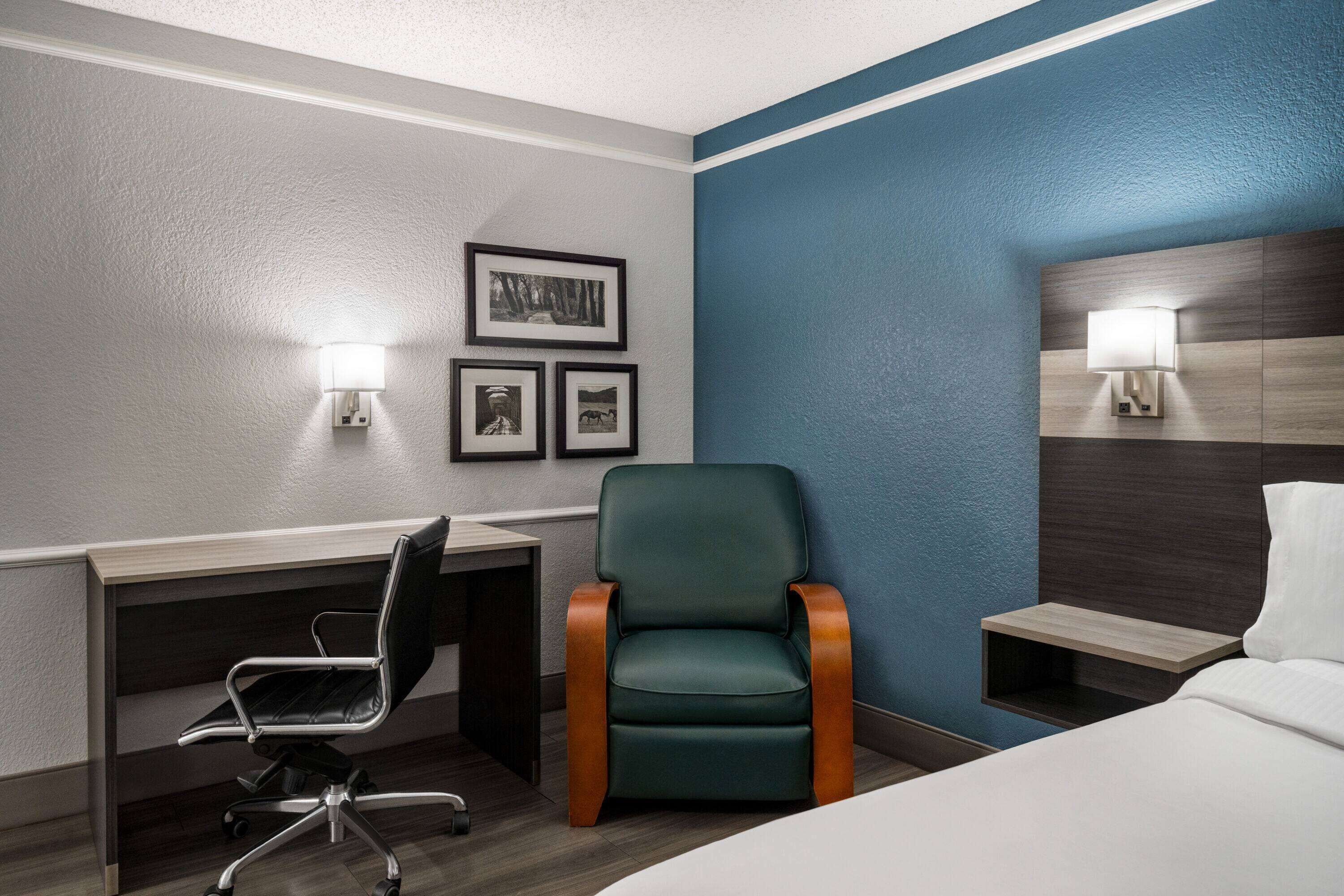 Een hotelkamer met een bureau, een groene fauteuil, een bed en een nachtkastje met een lamp.