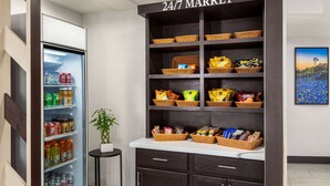 A 24/7 market with shelves stocked with snacks and beverages, a refrigerator with drinks, and a small table with a plant. - La Quinta Inn by Wyndham Victoria (Victoria)