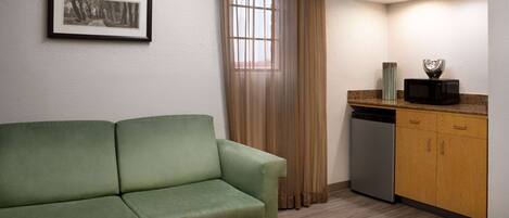A room with a green sofa, a kitchen area with a microwave, and a framed picture on the wall.