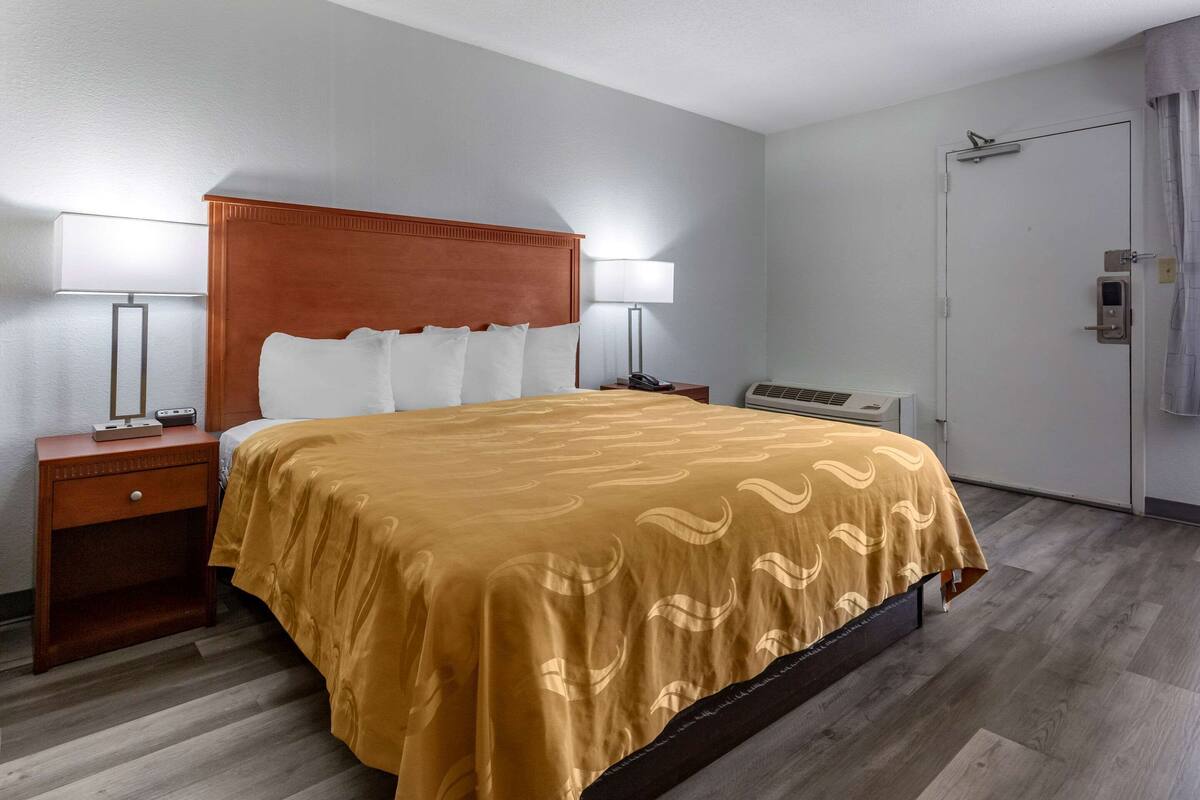 A hotel room with a bed, two bedside lamps, a nightstand, a heater, and a door with a curtain.